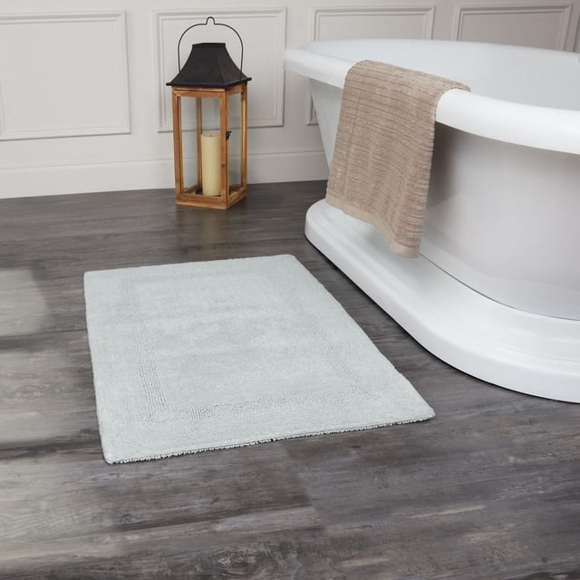 Fabstyles Basix Soft & Absorbent Reversible Cotton Bath Rug in light grey placed on dark wood floor next to white freestanding bathtub with beige towel draped over the tub edge and wooden lantern with candle in the background, made from 100% looped cotton for durability and absorbency, ideal for bathroom use.