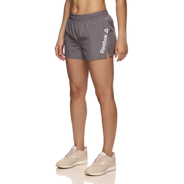 Reebok Women's Staple Running Short with Mesh Lining in grey polyester, featuring an elastic waistband with inside drawcord, side slit, and vertical Reebok Delta logo on the left leg, worn with beige running shoes on a white background, ideal for running, training, or casual wear.