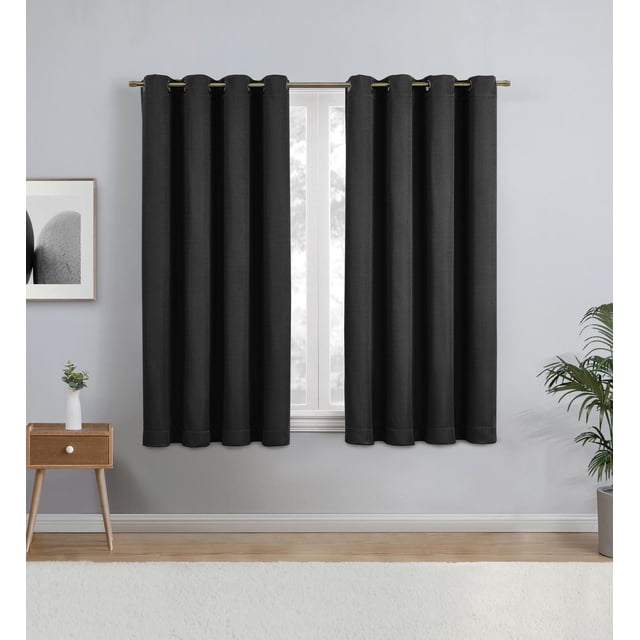Eclipse Heath 63" Grommet Blackout Window Curtains Panel in black hanging on a metal rod over a window with light grey walls. Made of textured polyester fabric with metal grommets, these curtains block light and reduce noise while providing privacy and energy savings. The setting includes a wooden side table with a small plant and a framed black and white photo. These blackout curtains are also suitable for creating a darkened space for media rooms or bedrooms.