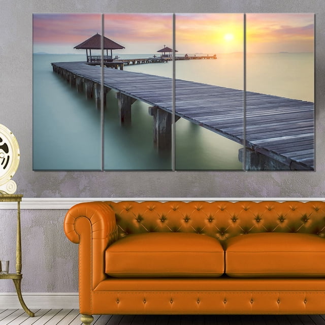 Design Art Wooden Sea Bridge And Sunset Seashore Photo Canvas Art Print depicting a four-panel gallery wrapped canvas of a wooden pier extending over calm water towards two thatched roof gazebos under a sunset sky, displayed above an orange tufted leather sofa, ideal for adding coastal charm to home décor.