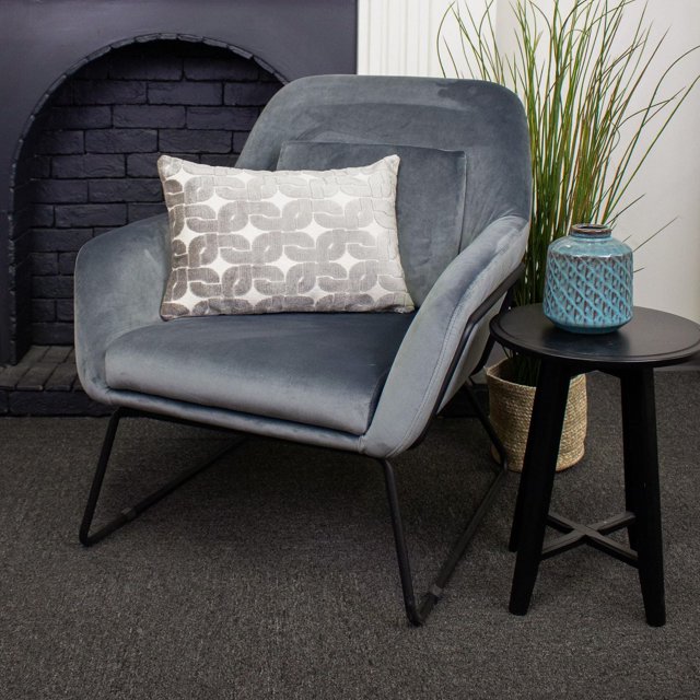 Haven Luxury Feather Filled Cushion measuring 12 by 20 inches with a light grey and beige geometric pattern, placed on a modern grey velvet armchair with black metal legs. This decorative throw pillow has a 100% polyester cover and is filled with premium white duck feathers. The setting includes a black side table with a textured blue ceramic vase and a tall green plant in a woven basket on a dark carpeted floor near a black brick fireplace.