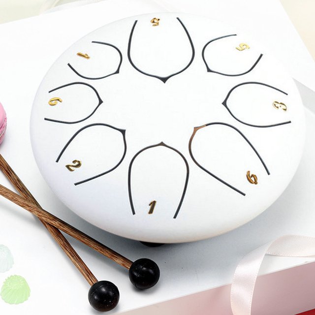 TopLLC Tambour de 6 Pouces à 8 Tons pour Adultes, Instrument de Musique à Main avec Fini Blanc et Numérotation Noire et Dorée sur Sa surface, Conçu pour la Méditation et la Détente. le Tambour Est Fait d'Acier au Carbone et Est Livré avec des Baguettes en Bois avec des Embouts en Caoutchouc Noirs. Il Est Présenté sur une surface Blanche avec des Accessoires Supplémentaires, Incluant un Ruban et des Articles Décoratifs à Proximité.