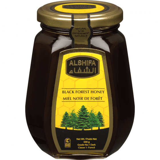 AlShifa Honey Black Forest in a dark glass jar with a gold lid, featuring a yellow label with green pine trees and bilingual English-French text, containing 500g of dark honey with rich forest floral flavor, natural enzymes, and immune support, suitable for toast or baking.