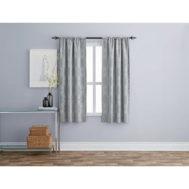 Eclipse Zana Trellis 63" Blackout Rod Pocket Window Panel in grey with a wavy interlocking geometric jacquard pattern, hanging on a black metal rod over a white-framed window in a minimalist room with light grey walls, wood flooring, and a glass console table with wicker baskets and decorative vases. The panel features premium thermal lining for blackout, privacy, energy savings, and noise reduction.