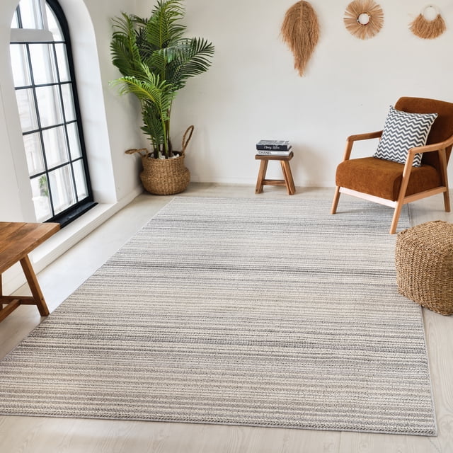 Rug Branch Jules Modern Indoor Area Rug in grey beige with subtle horizontal lines, on light wood floor in a bright room with a brown upholstered armchair with chevron pillow, woven basket planter with green palm leaves, small wooden stool with books, and natural fiber wall hangings.