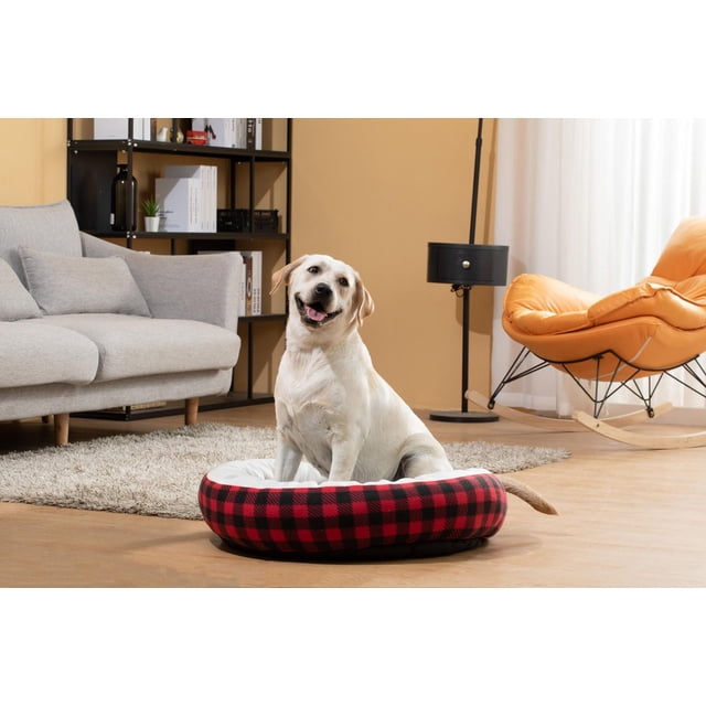 Pet Space Round Bed Red featuring a red and black checkered polyester fabric exterior with a soft white cushioned interior, placed on a hardwood floor in a living room with a gray sofa, black shelving unit, and orange rocking chair, ideal for dog comfort and often used as a cozy resting spot.