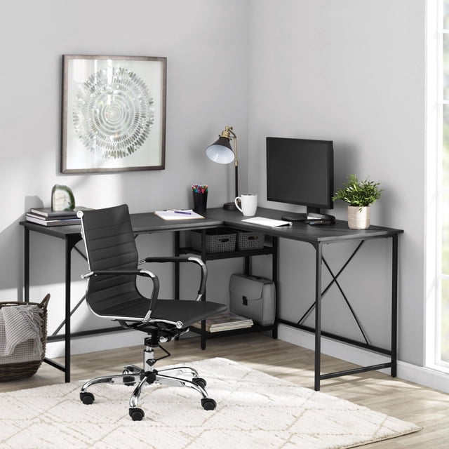 Mainstays Two-Way Convertible Desk with Lower Storage Shelf in black metal frame set up in an L-shape in a bright home office corner. The large desktop holds a computer monitor, desk lamp, mug, and potted plant. Two open shelves below store baskets and books for organized office furniture use. A black swivel chair with chrome base sits on a white patterned rug over light wood flooring, complemented by framed abstract wall art.