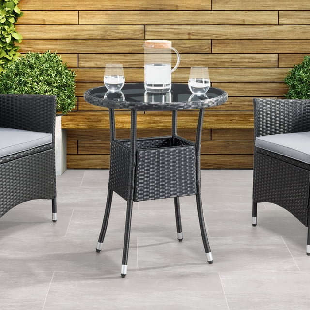CorLiving Parksville Black Steel and Resin Rattan Round Bistro Table with Glass Top featuring a black resin wicker frame and 5mm inset tinted black tempered glass top, supported by four powder-coated steel legs with metal wrapped cuffs and silver foot caps, set on a grey tiled patio with two matching black wicker chairs and green shrubs against a wooden plank wall, ideal for small outdoor spaces and casual alfresco dining.