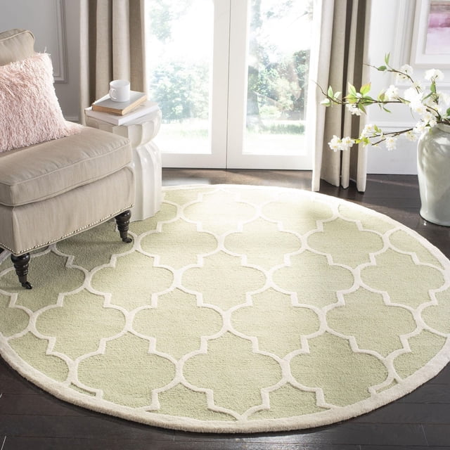 Safavieh Cambridge Kevin Geometric Area Rug, round with light green wool pile and white geometric trellis pattern, placed on dark hardwood floor in a bright room with beige armchair, pink pillow, white side table holding books and cup, and a vase of white blossoms by glass doors, ideal for adding modern American style to home decor.
