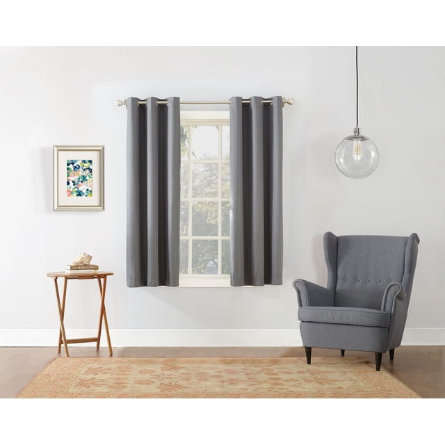 MAINSTAYS Textured Thermal-Lined Grommet Window Curtains Panel in dark grey with woven texture and thermal blackout lining hanging on a silver metal rod over a window in a minimalist room with white walls, hardwood floor, a grey wingback armchair, wooden side table with books and shell, framed abstract floral print, and clear glass pendant light. The thermal lining reduces energy loss and noise, enhancing room comfort.