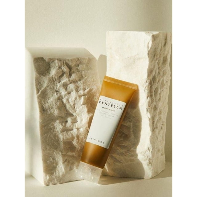 SKIN1004 Madagascar Centella Ampoule Foam 125ml tube with amber and white packaging, leaning against two textured beige stone blocks on a neutral background, a hydrating and calming daily facial cleanser enriched with Centella Asiatica extract.