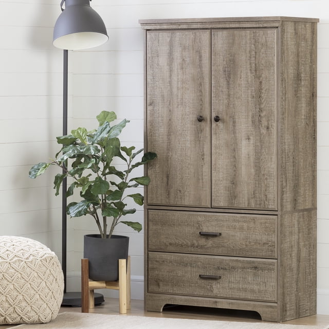 South Shore Versa 2-Door Armoire with Drawers in weathered oak finish, showing two doors with round knobs above two spacious drawers with rectangular handles, standing on a light wood floor next to a black floor lamp and a green potted plant in a room with white shiplap walls, suitable for bedroom or living room storage.