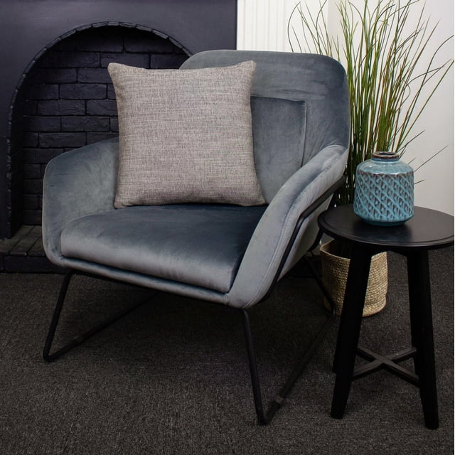 Remy Luxury Cushion grey decorative throw pillow made of 100% polyester on a modern grey velvet armchair with black metal legs, beside a black round side table holding a blue textured ceramic vase, set in a room with a black brick fireplace and tall green grass in a woven basket, suitable for enhancing home décor comfort and style.