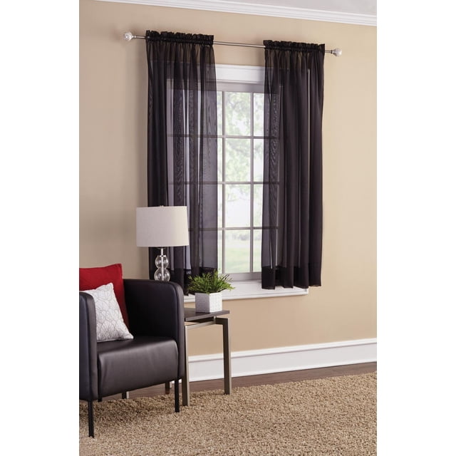 SUN TEXTILE CURTAIN à Passe-Tringle en VOILE Noir, Tissu VOILE Transparent Suspendu sur une Tringle Argentée au-Dessus d'Une Fenêtre Encadrée Blanche dans une Pièce beige, Filtrant Doucement la Lumière Naturelle et Améliorant l'Intimité, Idéal pour Superposer avec d'Autres Rideaux Transparents, Montré avec une Chaise en Cuir Noire avec des Coussins Rouges et Blancs, une table d'Appoint avec une Lampe en Verre et une Petite Plante Verte, sur un Plancher Tapisé beige.