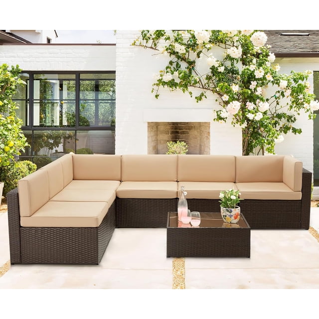 AECOJOY 7 PCS Patio Conversation Set with brown PE rattan wicker and beige cushions, arranged in an L-shape with a glass-top coffee table. This modular outdoor patio furniture set is ideal for gardens, balconies, or poolside spaces, shown in a garden setting with a white brick fireplace and greenery.