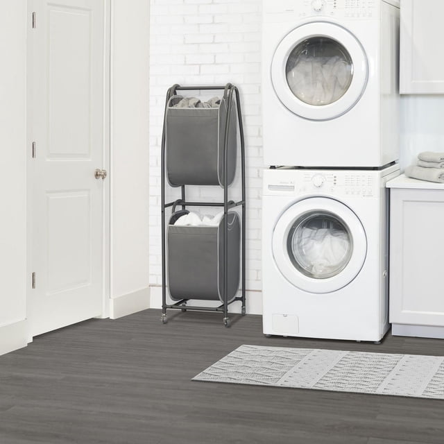 neatfreak! 2-Tier Rolling Vertical Laundry Sorter with Hamper-Totes and EVERFRESH® Odor Control in charcoal grey, featuring a sturdy metal A-frame with two collapsible fabric totes with C-shaped foam handles on casters, placed beside a stacked white washer and dryer in a laundry room with white brick walls and dark grey flooring, ideal for maximizing vertical space in compact laundry areas.
