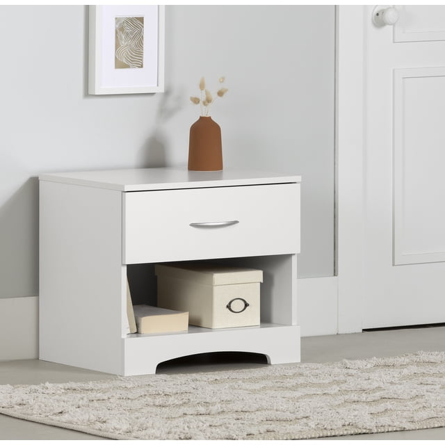 South Shore Step One 1-Drawer Nightstand in pure white with a modern cutout kickplate, featuring a metal handle drawer and open storage space holding a beige box and books, placed on a light textured rug against a grey wall with framed abstract art and a brown vase on top, suitable for bedside storage and organization.