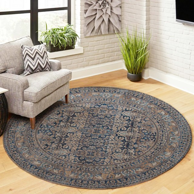 ECARPET Ariana Traditional Area Rug in a round shape with blue and beige repeating micro patterns, placed on a light hardwood floor in a bright living room corner featuring a grey armchair with a chevron pillow, green plants, and white brick walls, ideal for grounding neutral and wood-toned spaces.