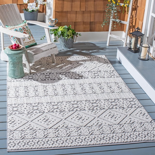 SAFAVIEH Cabana Allycia Tapis Géométrique Intérieur/extérieur sur un Plancher de Porche en Bois Gris, Présentant un Motif Géométrique Texturé Crème et Gris avec des motifs de Losanges et Floraux. Assorti à une Chaise Adirondack Blanche avec un Coussin à motifs, une table d'Appoint en Céramique Verte avec des Pommes Rouges et des Plantes en Pot. des Lanternes et des Bougies sur une Marche Grise Surélevée Complètent le Décor Extérieur. Ce Tapis Extérieur durable Est Idéal pour les Espaces Intérieurs Ou Extérieurs Occupés et Est Souvent Utilisé pour Ajouter du Charme de Villégiature Tropicale aux Terrasses