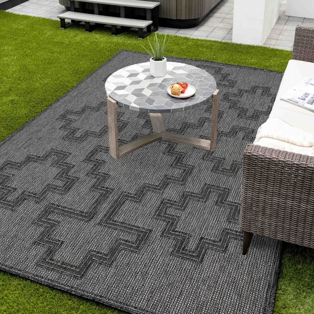 ECARPET Esina Transitional Area Rug in dark grey with textured geometric cross patterns, placed outdoors on green grass under a round mosaic tile coffee table with wooden legs, beside a wicker sofa with white cushions and a folded blanket, ideal for patio or indoor use.