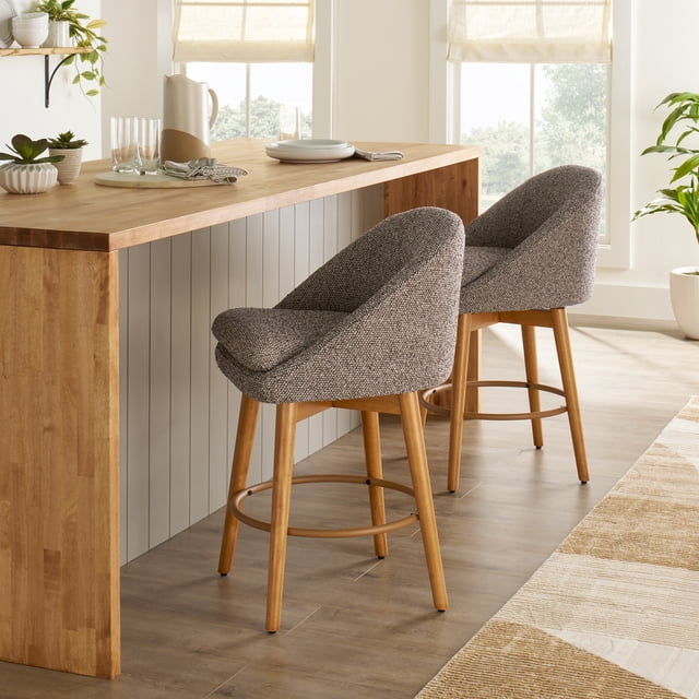 Better Homes & Gardens Mira Counter Stool 2 Pack with two truffle brown textured fabric stools featuring light honey solid wood bases and 360-degree swivel rings, placed at a wooden kitchen island in a bright room with neutral decor, suitable for kitchen seating or casual dining.