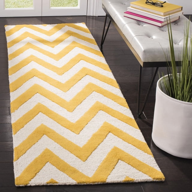 Safavieh Cambridge Kaitlyn Zig Zag Stripes Area Rug with yellow and white zig zag stripes in hand-tufted wool, placed on dark hardwood floor beside a white tufted bench with books and glasses, and a white planter with green grass-like plant, ideal for adding texture and modern American style to home or office decor.