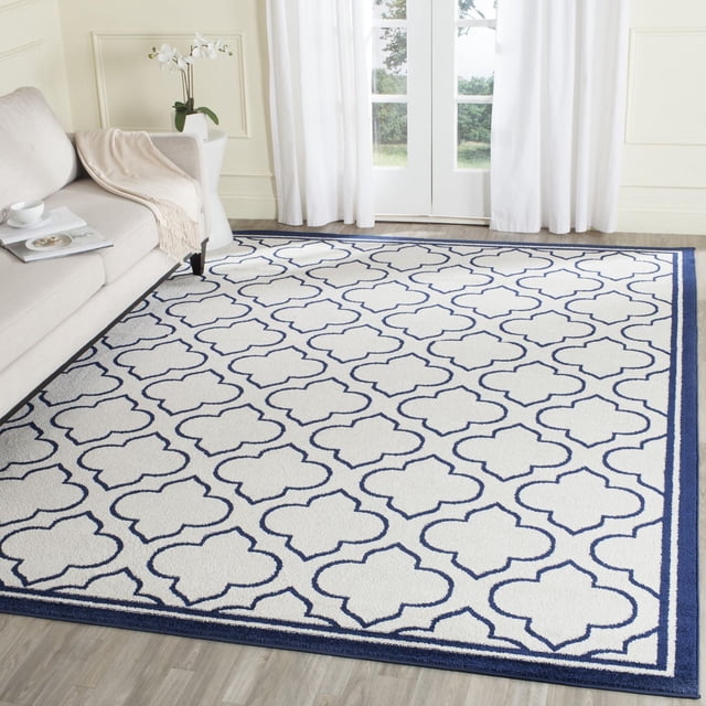 Safavieh Amherst Wilson Tapis Géométrique pour Aire avec Fond Blanc et Motif de Trèfle Bleu Marine, Bordé en Bleu Marine, construction en Polypropylène Tissée Mécaniquement Idéale pour un Fort Achalandage Intérieur Ou Extérieur, Exposé sur un Plancher de Bois Clair à Côté d'Un sofa beige et de Rideaux Blancs.