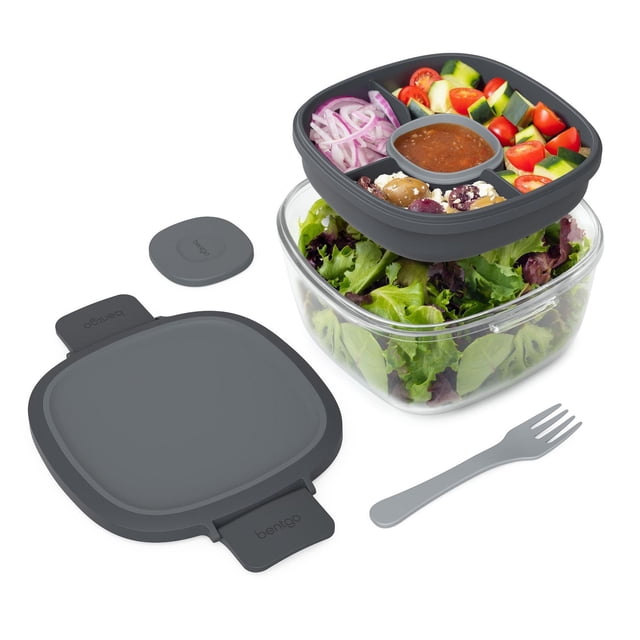 Bentgo Glass All-in-One Salad Container in dark gray with a clear glass bowl holding mixed greens, a removable divided tray containing cherry tomatoes, cucumber pieces, sliced red onions, olives with feta cheese, and a leak-proof dressing container. Includes a dark gray lid and fork for mess-free salad transport and mixing.
