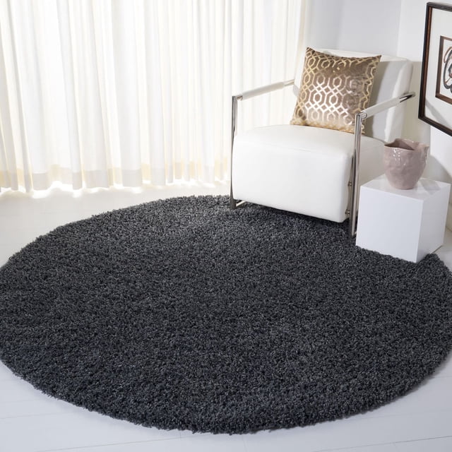 Safavieh Venus Elijah Solid Plush Shag Area Rug, round with a dense 1.77-inch medium pile in dark charcoal grey, placed on a light floor beside a white modern armchair with chrome arms and a gold geometric pillow, next to a white cube side table with a mauve ceramic vase, in a bright room with sheer white curtains, ideal for adding warm texture and dimension to contemporary living spaces.
