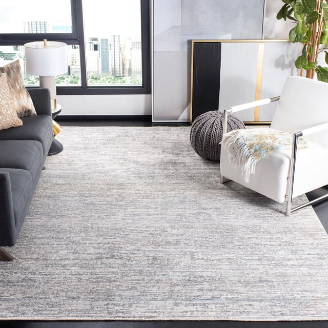 SAFAVIEH Dream Ethelyn Abstract Overdyed Area Rug, an 8'x10' durable accent rug with a muted abstract pattern in grey and beige, shown in a modern living room with a dark grey sofa, white armchair with a fringed throw, knitted grey pouf, and contemporary artwork near a large window overlooking a cityscape. Suitable for adding subtle texture and warmth to home décor.