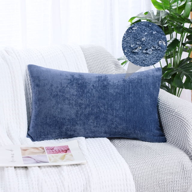 PiccoCasa Soft Water Repellent Throw Pillow Covers Navy Blue 12x20 Inch displayed on a white and gray textured armchair. The decorative pillow cover is made of chenille material with a soft, thick texture and features a solid navy blue color. A close-up inset highlights the water-repellent coating, showing water droplets on the fabric. The setting includes a bright room with natural light and green plants in the background.