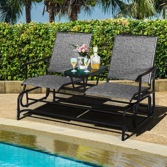 Costway 2 Person Outdoor Patio Double Glider Chair Loveseat Rocking w/Center Table Gray. A two-seater outdoor glider bench with a solid steel frame and gray weather-resistant fabric. Includes a tempered glass center table for drinks or decorations, ergonomic armrests, and anti-slip foot pads. Ideal for outdoor seating in patios, gardens, or poolside areas, with lush greenery in the background.