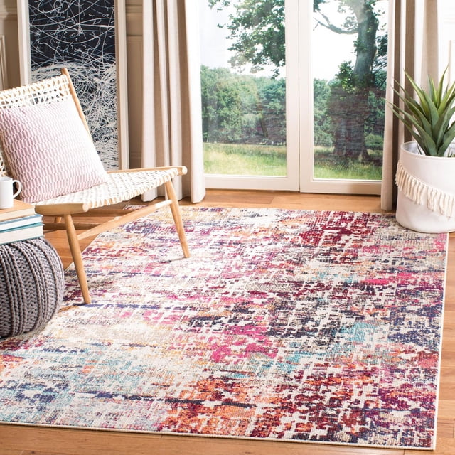 Safavieh Madison Joella Vintage Abstract Area Rug featuring a multicolored abstract pattern in red, pink, blue, orange, and cream on wooden flooring in a bright room. This transitional accent rug lies beneath a woven chair with a pink cushion, a gray knitted pouf, and a white fringed planter near glass doors with a green outdoor view. Often used to enhance modern and contemporary home decor styles.