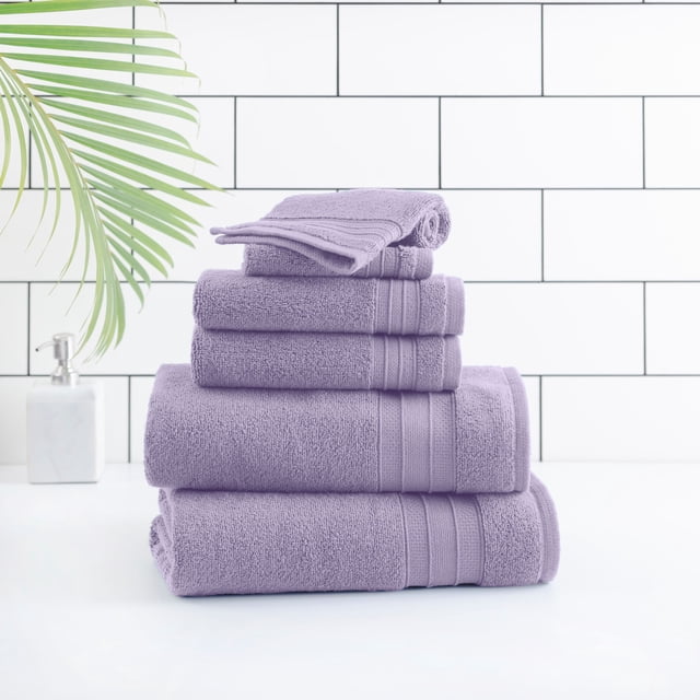 hometrends Solid Bath Towel 6-Piece Set in purple stacked on a white surface with a white tiled wall background, made from 100% cotton with absorbent terry texture and ribbed bands, shown with a green palm leaf and white soap dispenser, suitable for a spa-like bathroom and often used as a soft wrap for delicate items.