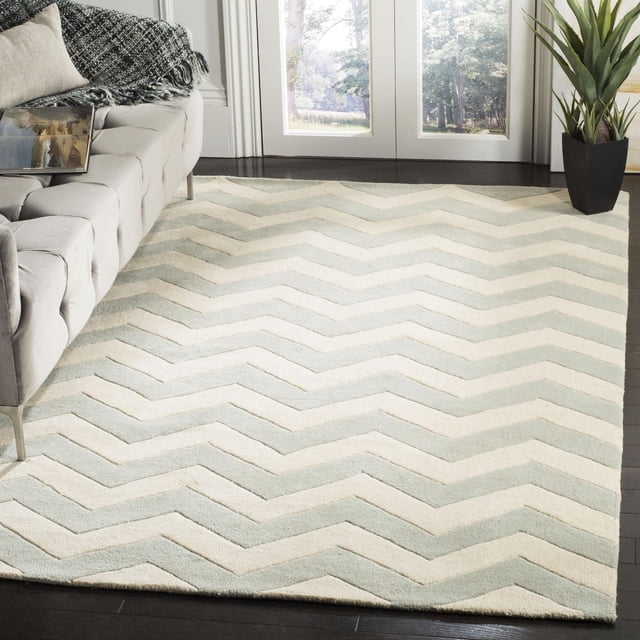 Safavieh Chatham Jake Zigzag Stripes Area Rug featuring hand-tufted wool with a medium pile and a chevron zigzag pattern in cream and light grey. Displayed on dark hardwood flooring beside a grey tufted sofa and near glass doors with a view of trees, ideal for adding softness and contemporary style to living spaces.