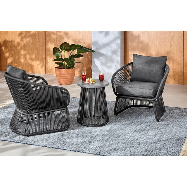 HOMETRENDS Corday Steel 3-Piece Outdoor Chat Set with Cushions, Black, featuring two black handwoven resin wicker chairs with dark grey UV- and fade-resistant cushions and a black steel side table with a hand-painted faux stone finish, arranged on a blue patterned outdoor rug with a terracotta potted plant in the background, ideal for casual outdoor seating and small gatherings.