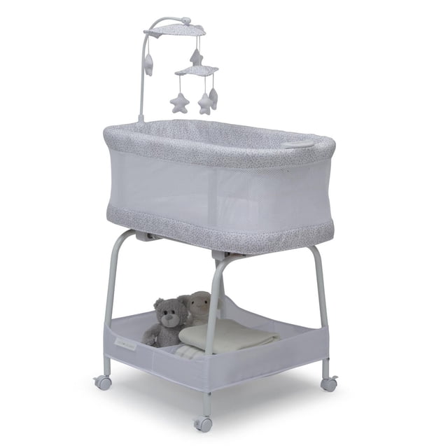 Delta Children Sweet Breeze Bassinet with Airflow Mesh in light grey with small dot pattern, featuring breathable mesh sides for airflow and visibility, an overhead toy arm with plush stars, a large storage basket underneath holding two stuffed animals and folded blankets, and four locking caster wheels for easy movement.