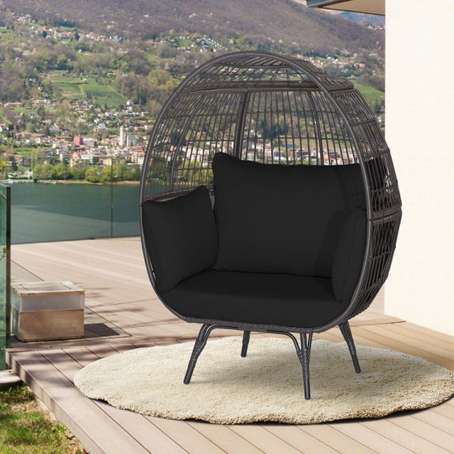 Costway Patio Oversized Rattan Wicker Egg Chair Lounge Basket with black cushions, featuring a mix brown hand-woven PE wicker frame and a spacious egg-shaped design. Ideal for outdoor seating or patio furniture, the chair is placed on a wooden patio with a scenic view of a lake and hills in the background.