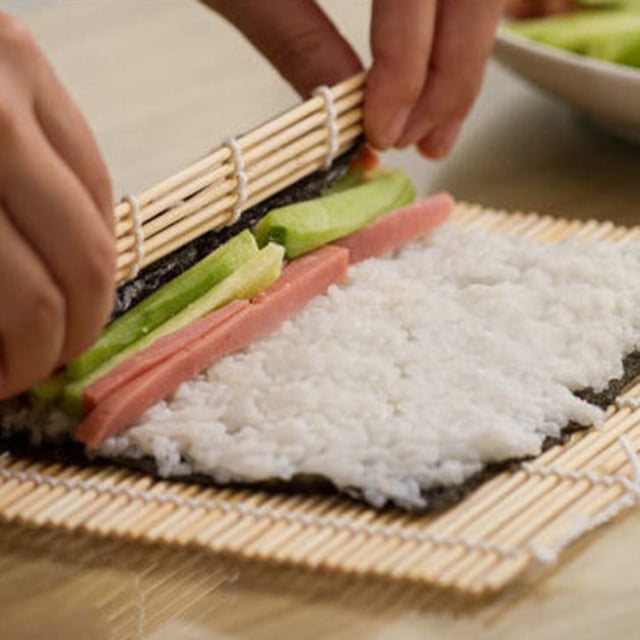 HEALEEP Sushi Making Kit includes a bamboo sushi rolling mat and rice paddle, shown in use with hands rolling sushi. The mat holds rice, seaweed, and fillings like cucumber and ham, creating a traditional sushi roll. Ideal for beginners and home cooks, this kit simplifies sushi preparation.