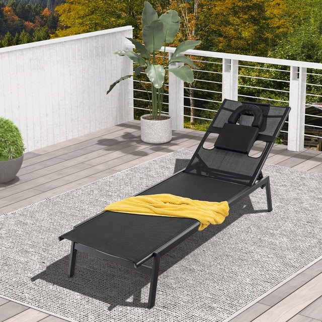 Gymax Patio Sunbathing Lounge Chair with face hole and detachable head pillows, shown in black with a metal frame and rip-resistant fabric. The chair features a 5-position adjustable backrest, a U-shaped pillow, and a headrest pillow secured with hook and loop fasteners. It is placed on a patio with a grey rug, surrounded by greenery and a white railing.
