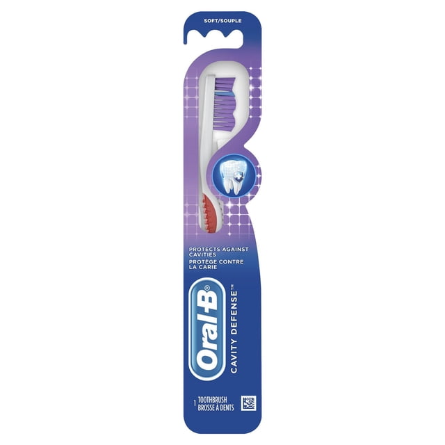 Oral-B Cavity Defense Toothbrush, Soft, in blue and purple packaging featuring a white toothbrush with purple and blue wave-trimmed soft bristles and a red ergonomic grip handle, designed to remove plaque and protect against cavities.