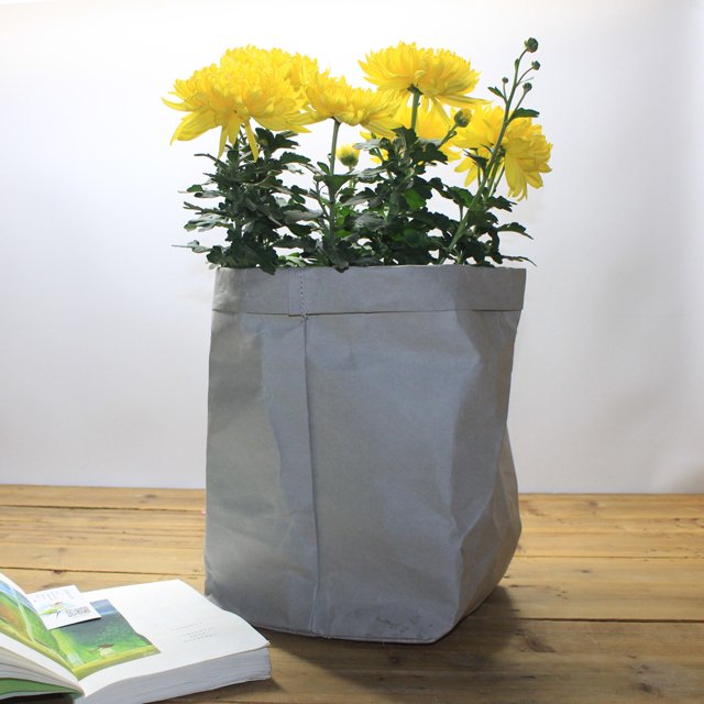 White Kraft Paper Bags Washable Kraft Paper Bag Plant Flowers Pots Multifunction Home Storage Bag Reuse. A grey washable kraft paper bag with a rectangular shape, holding vibrant yellow flowers. The bag is made from tear-resistant, environmentally friendly kraft paper and features a shimmering finish. It is placed on a wooden surface with an open book nearby, showcasing its versatility as a plant pot cover or storage solution.