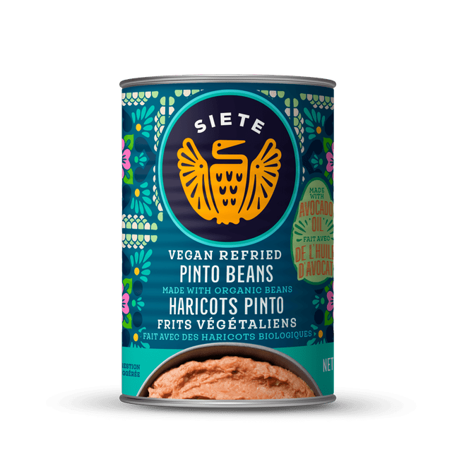 Siete Vegan Refried Pinto Beans canned food with teal floral label and orange bird logo, made with organic beans and avocado oil, showing smooth refried beans at the bottom, ideal as a plant-based protein pantry staple.