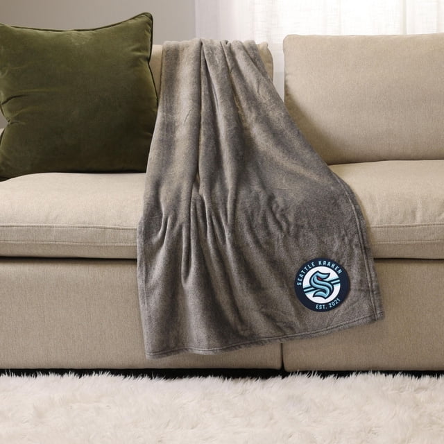 NHL, NBA, MLB Grey Sports Throw Blanket, 50" x 60", draped over a beige sofa with a green cushion, featuring a Seattle Kraken logo patch in blue and white near the bottom corner. This plush 100% polyester throw blanket provides warmth and softness, suitable for cozy indoor lounging or outdoor use.