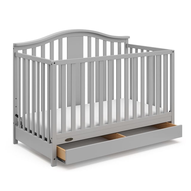 Graco Solano 4-in-1 Convertible Crib with Drawer in light grey finish, front 3/4 view showing slatted sides, curved headboard, and open full-size storage drawer with two compartments for nursery essentials, designed with adjustable mattress support for growing babies.