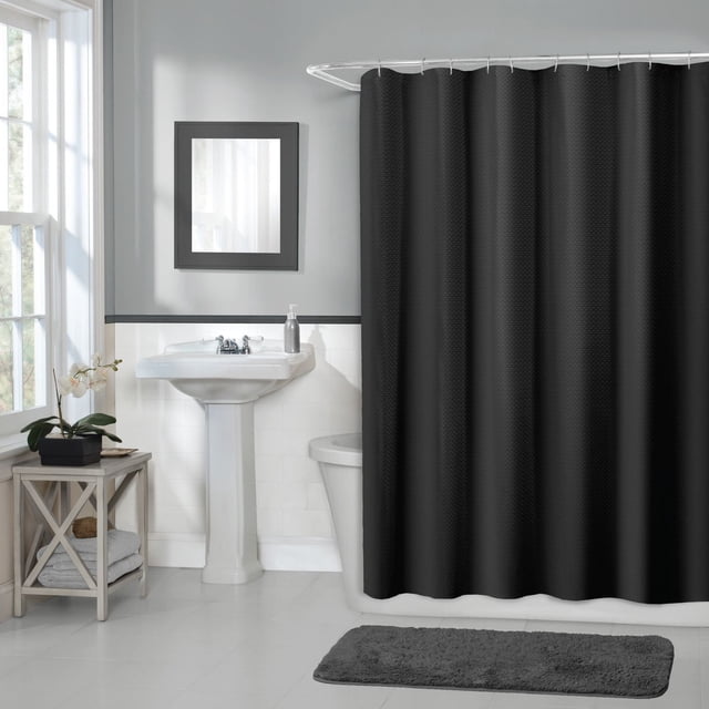 Hometrends Waffle Fabric Shower Curtain, 70 Inches x 72 Inches, Black, displayed in a bathroom with white tile and a pedestal sink. The fabric curtain features a solid waffle texture and a buttonhole top for easy hanging. A dark gray bath mat and a wooden side table with towels and a potted orchid complete the scene. The curtain is machine washable and fits standard tubs.