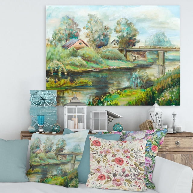 Designart Bridge On The River In Rustic Landscape Canvas Wall Art featuring a gallery-wrapped premium cotton canvas print of a tranquil river scene with a wooden bridge, rustic houses, and lush greenery, displayed above a wooden console with matching decorative pillows and farmhouse-style lanterns, suitable for rustic home décor.