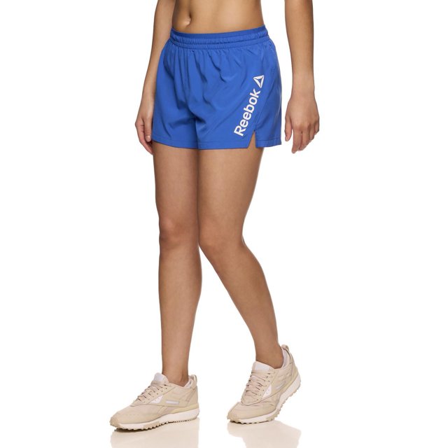 Reebok Women's Staple Running Short with Mesh Lining in blue, shown on a model from waist to mid-thigh, made of lightweight 100% polyester with elastic waistband and inside drawcord, side slit, and white Reebok Delta logo on the left leg, ideal for running, training, or casual wear.