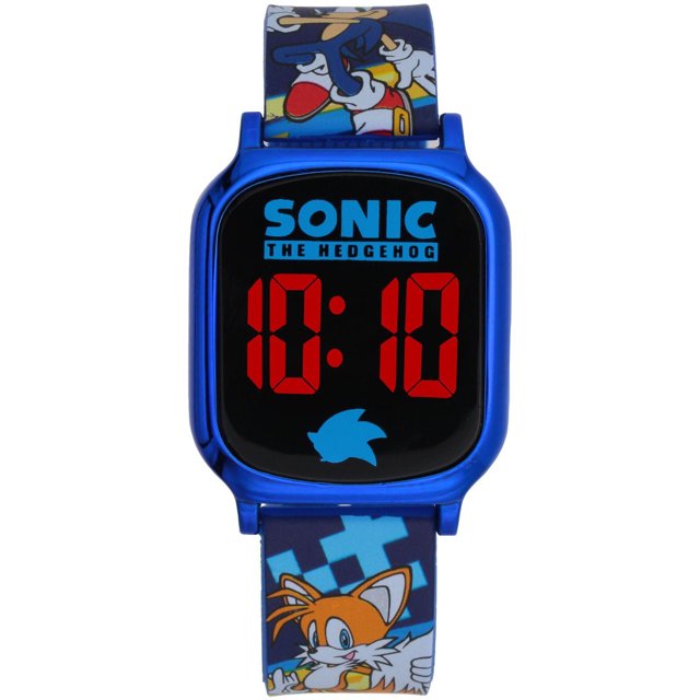 Sonic the Hedgehog Kids’ Digital LED Watch with a blue plastic case and red digital display showing 10:10. The watch face features the Sonic the Hedgehog logo and a blue Sonic silhouette. The blue silicone strap displays colorful Sonic and Tails graphics on a pixelated background with a metal buckle, suitable for children aged 6 and up.