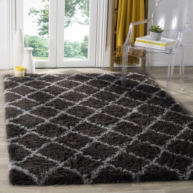 Safavieh Indie Careen Geometric Shag Area Rug featuring a plush dark gray shag pile with a lighter gray diamond geometric pattern, placed on light wood flooring near glass doors, complemented by a clear acrylic chair with books and a small succulent, suitable for enhancing texture and comfort in accent rug spaces.